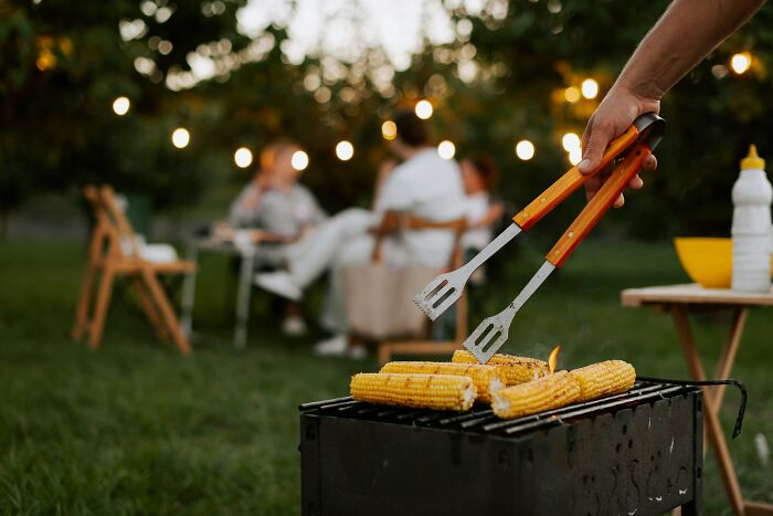 A hand holding tongs to grill corn on a barbecue at an outdoor gathering.