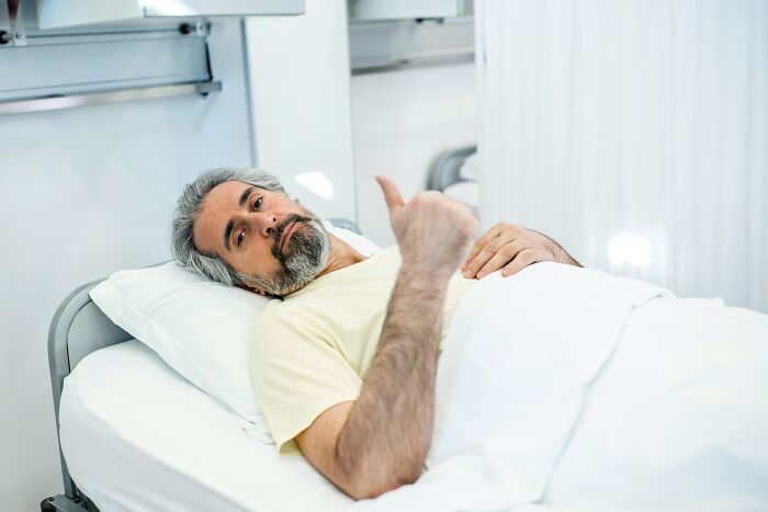 A medical patient lies in a hospital bed while showing a thumbs-up sign.
