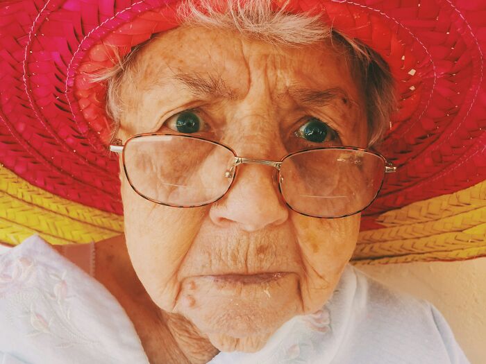 An elderly woman wearing glasses and a large hat looking closely, showing funny patient stories