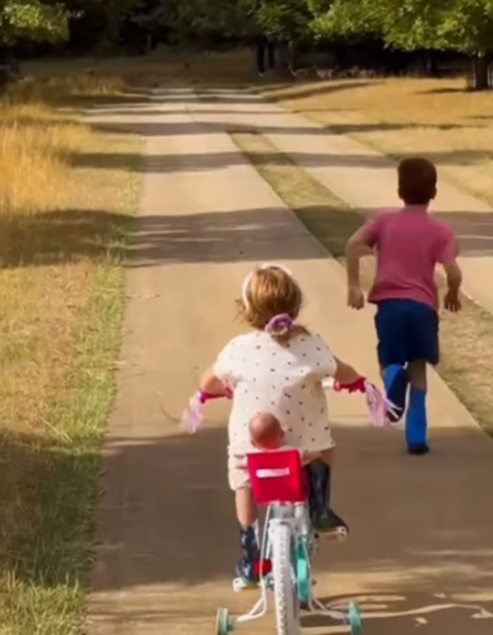 Two children run down a paved path, one on a small bicycle and the other running behind. Meghan Markle's post. Two children run down a paved path, one on a small bicycle and the other running behind. Meghan Markle's post.
