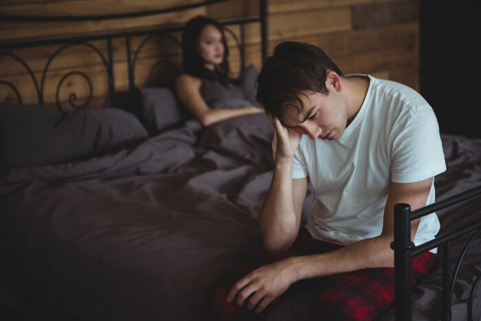Upset man sitting on bed with woman in background during relationship conflict