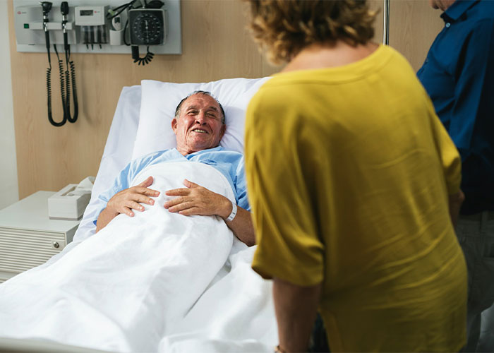A man smiling in a hospital bed with visitors, highlighting why MIL is angry.