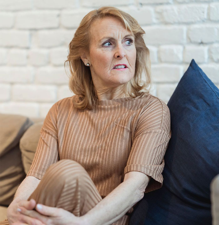 A woman sits on a couch with a shocked expression, learning why her MIL is angry.