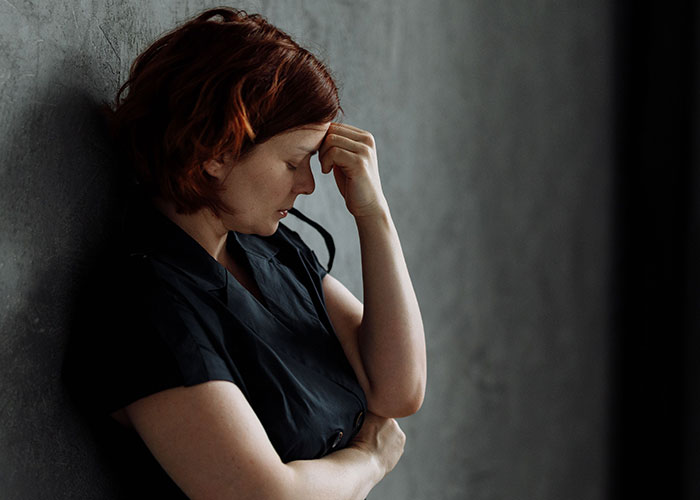 Stressed woman in black shirt leaning against wall with hand on forehead, reflecting deep worry