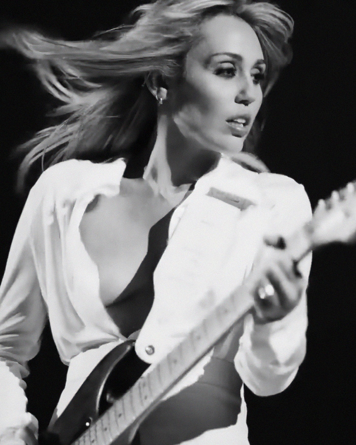 Miley Cyrus, with flowing hair, plays guitar on stage, her name change sparking fury after Dolly Parton's passing.
