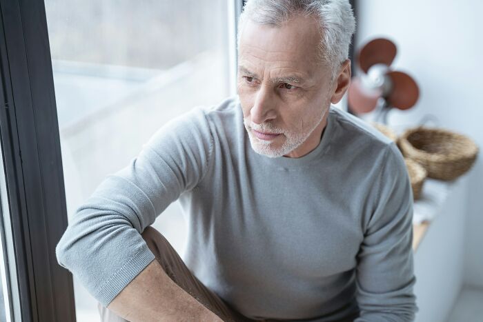 Elderly man looking out a window, contemplating midlife crisis.