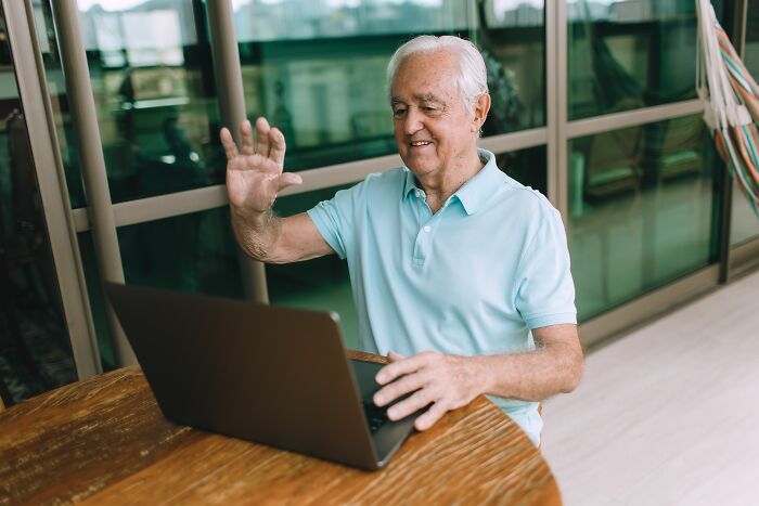 A senior man waves to his laptop screen, likely experiencing a midlife crisis with new technology.