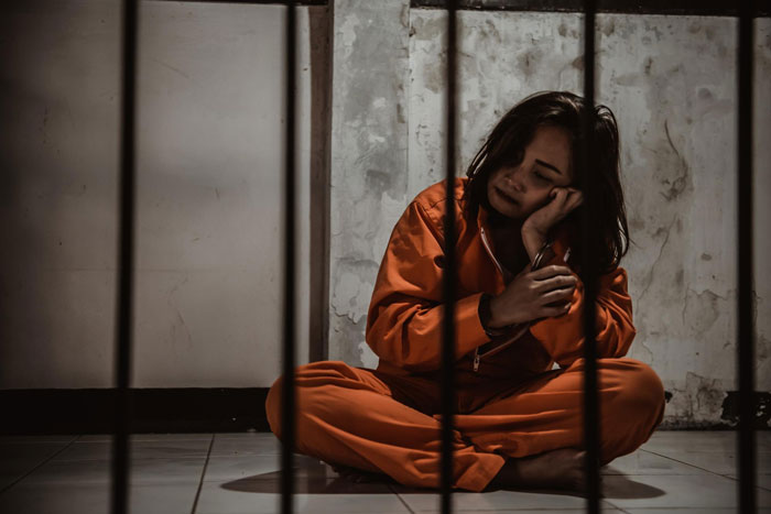 Woman in an orange jumpsuit behind bars, looking sad and holding her hand to her cheek after a prison break. Woman in an orange jumpsuit behind bars, looking sad and holding her hand to her cheek after a prison break.