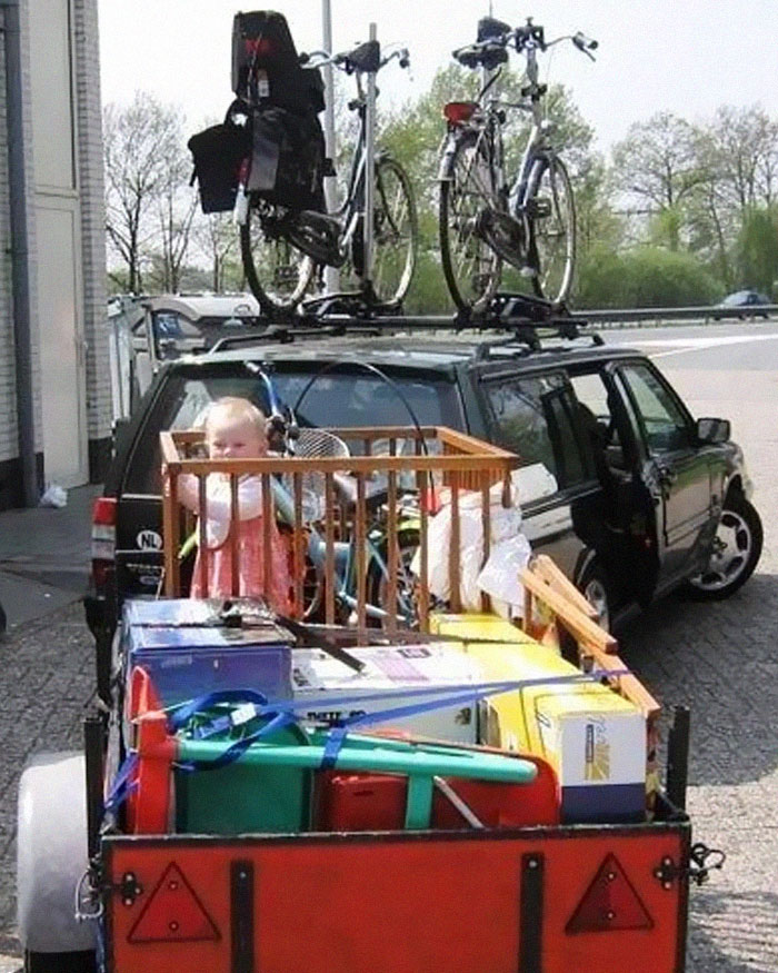 Child in crib and household items loaded on trailer behind car with bikes on roof, a moving fails setup.