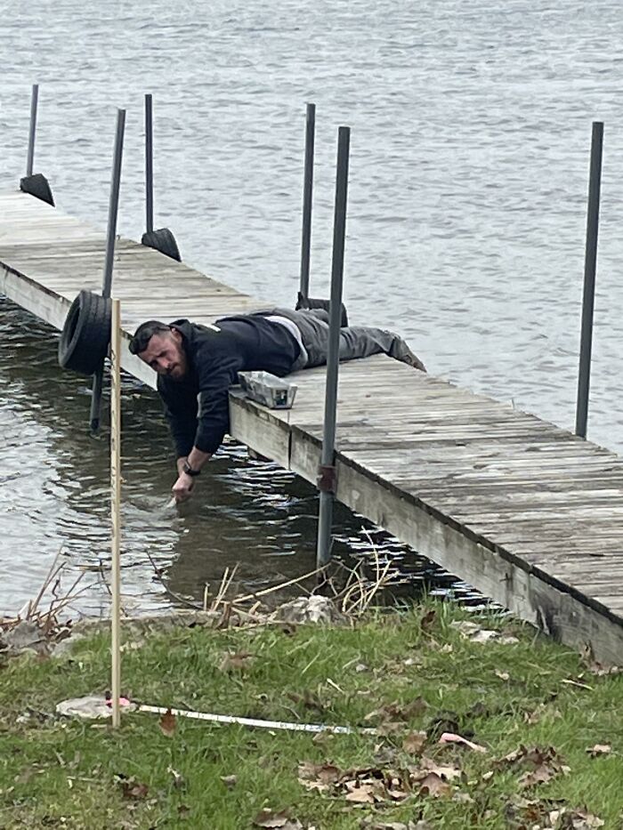 A man lying on a wooden dock reaches into the water, a confusing perspective making him look tiny.