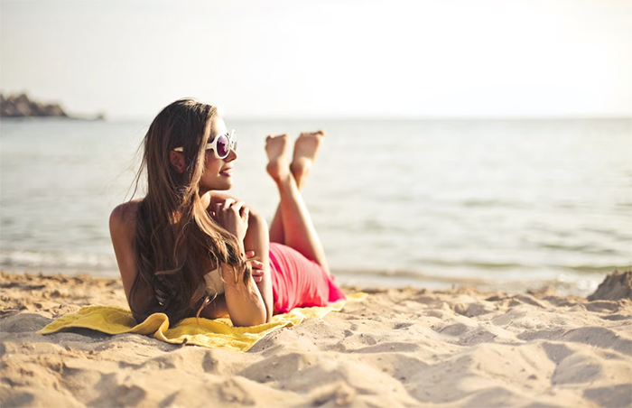 A woman in a red bikini top and sunglasses lies on a yellow towel on a sandy beach, facing the ocean. A woman in a red bikini top and sunglasses lies on a yellow towel on a sandy beach, facing the ocean.