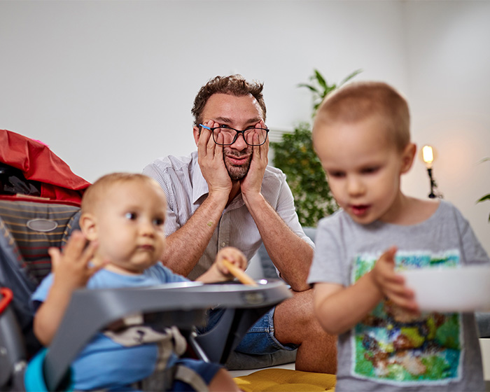 Tired dad looking overwhelmed while two young children are active, fueling the viral debate on parenting.