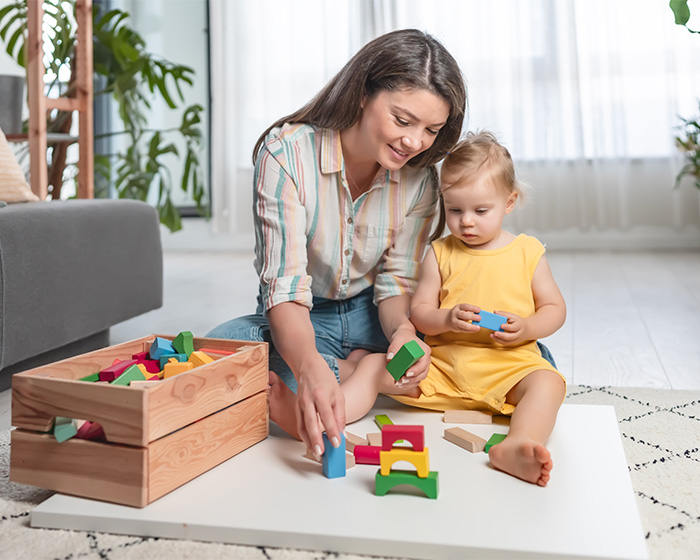Mother and child playing with building blocks on the floor, sparking debate about parenting and nursery care.