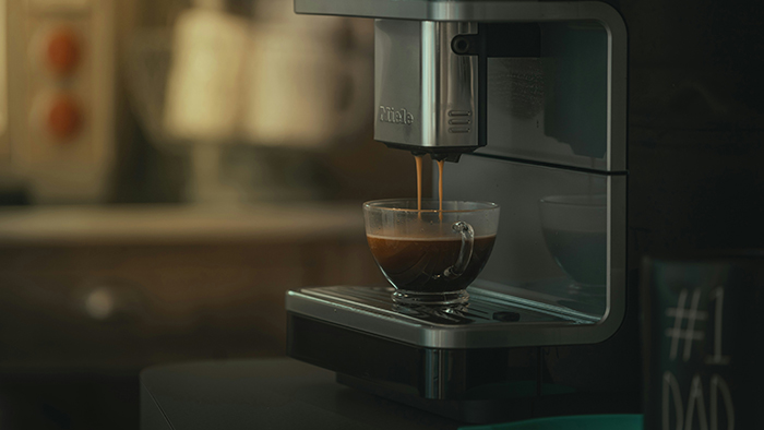 Espresso brewing from a modern coffee machine into a clear glass cup in an office setting.