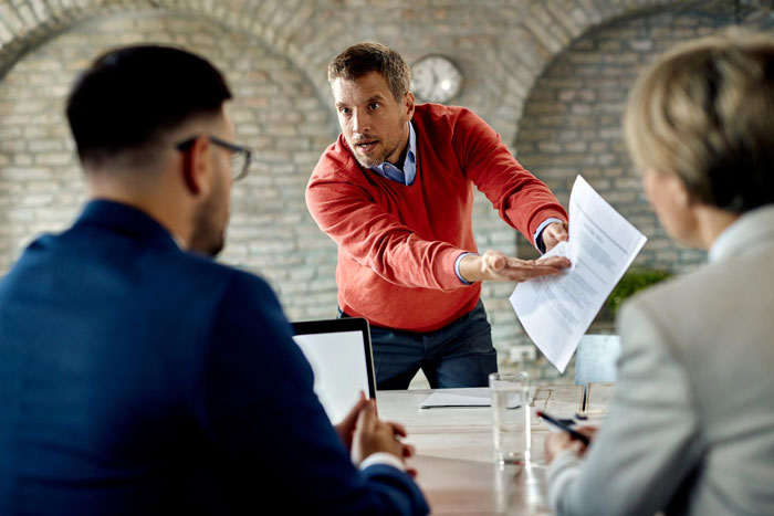 A frustrated boss pointing at documents in a meeting, expecting an employee at HQ after six years remote. A frustrated boss pointing at documents in a meeting, expecting an employee at HQ after six years remote.