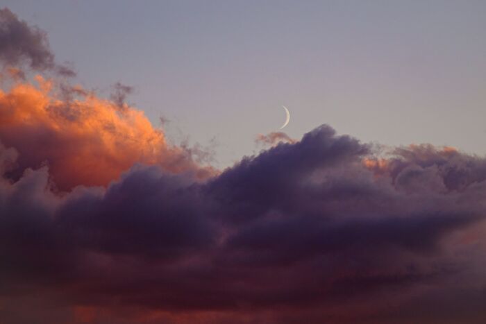 A crescent moon visible through colorful, dramatic clouds at dusk, offering cool facts about the universe's celestial bodies.