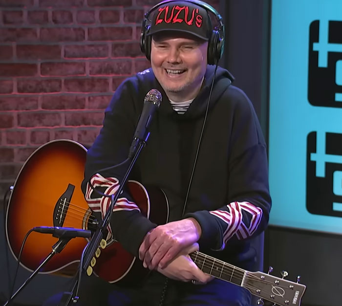 Billy Corgan, one of the famous celebrities, smiling with his acoustic guitar in a casual studio setting