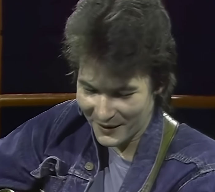 A young musician, representing celebrities visiting ordinary people, smiling while playing an acoustic guitar