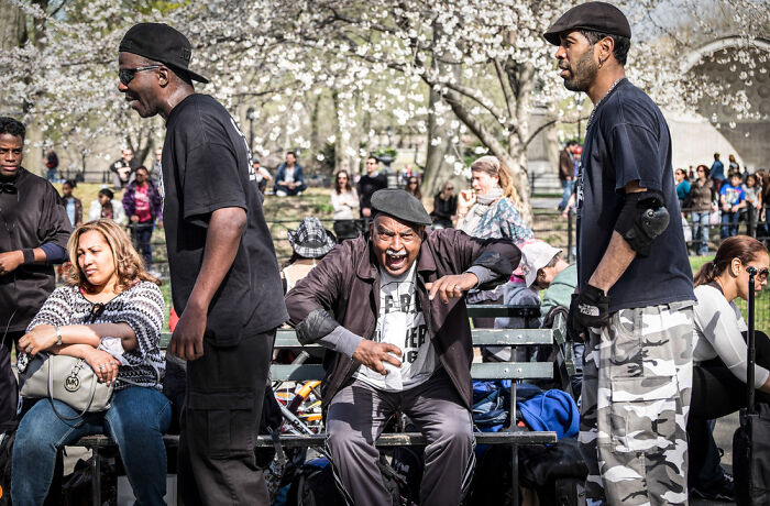 A street photographer captures an odd coincidence: a man in a black hat making a funny face in a park with people.