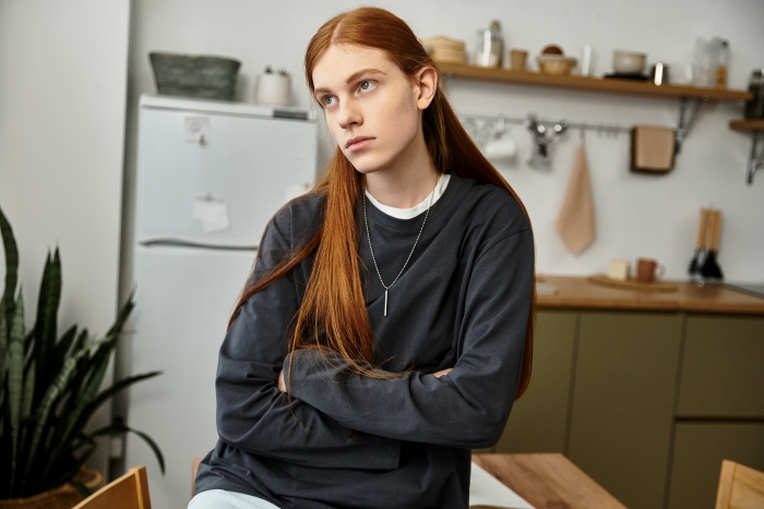 An upset stepdaughter with long red hair sits with arms crossed in her kitchen looking away.