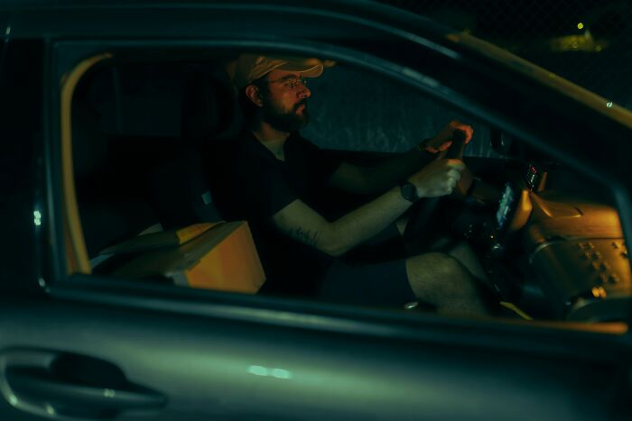 A man with a beard and cap driving a car at night, perhaps relying on his 6th sense about a situation.