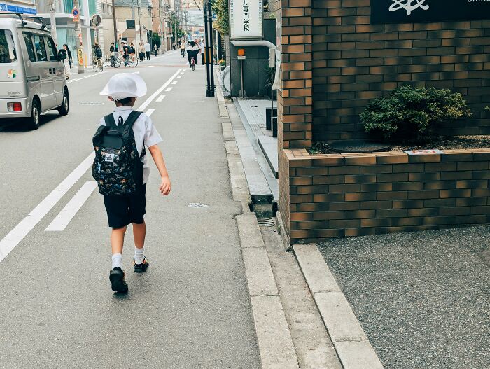 A child in a white hat and backpack walking on a street, perhaps their 6th sense is developing.