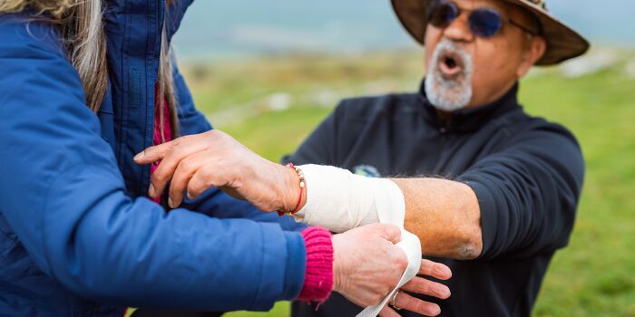 A woman bandaging a man's arm, who appears to be reacting to pain with his mouth open.
