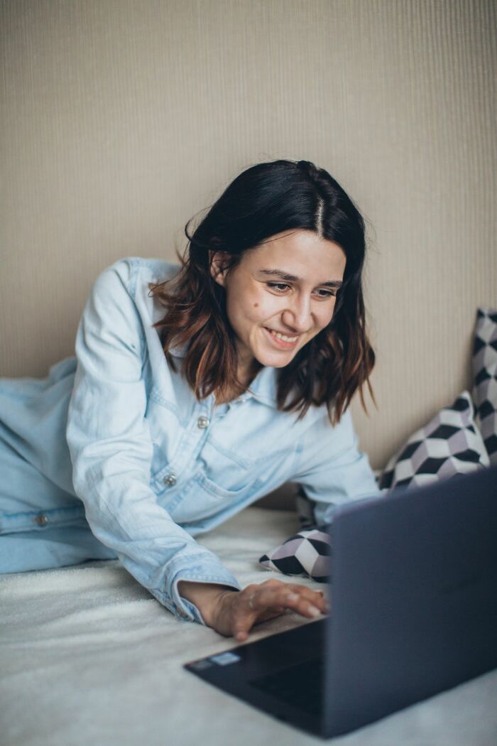 A smiling woman lying on a bed using a laptop, showing a moment of peace after relationships have ended.