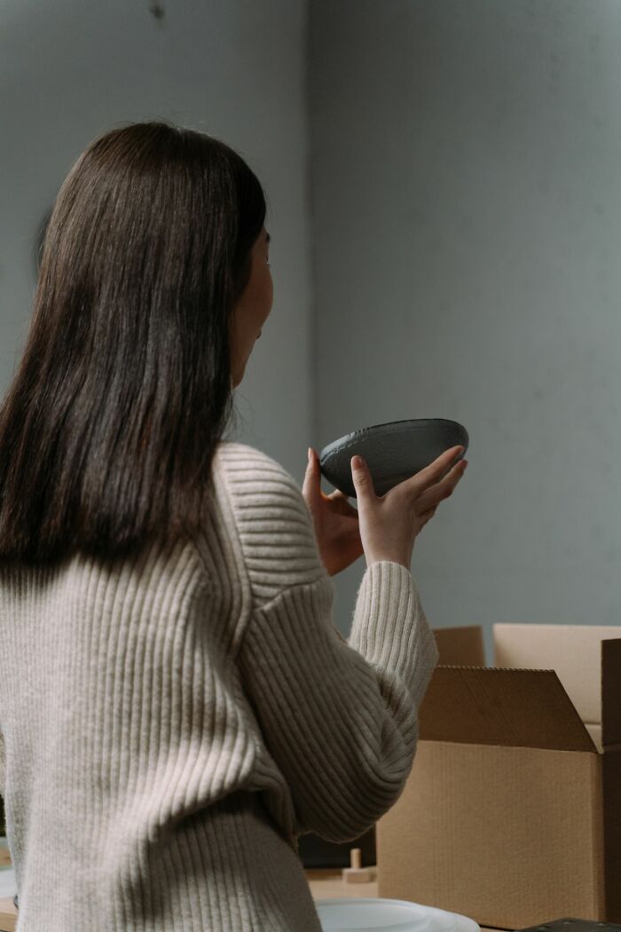 A woman wearing a beige sweater holds a small dark dish, looking at it intently, perhaps reflecting on relationships.