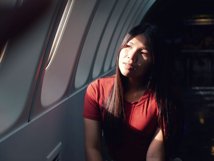 A young woman in a red shirt looking out the window while sitting next to a plane seatmate on a flight.