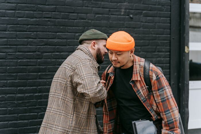 Two men in beanies whisper closely, one speaking into the other's ear, depicting an overheard moment.