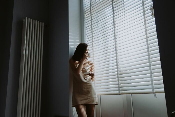A woman looking out a window with closed blinds, considering safety tips for women living alone.