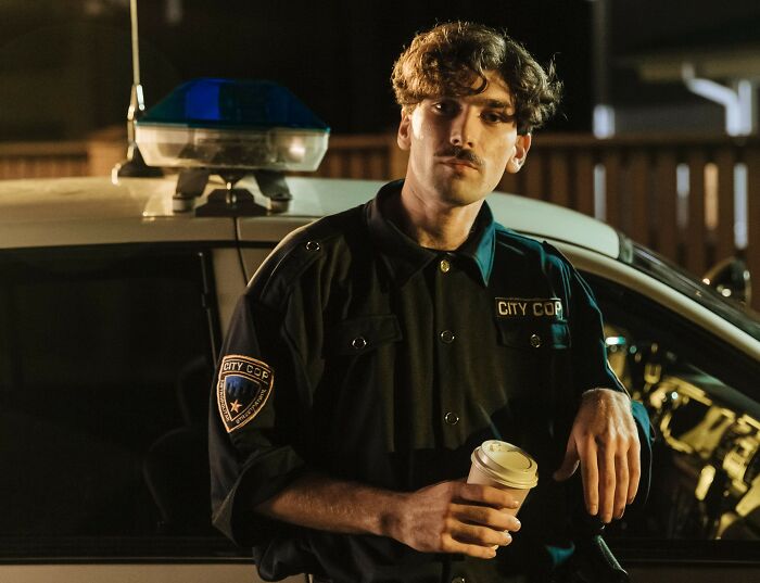 City cop in uniform holding coffee cup beside police car at night