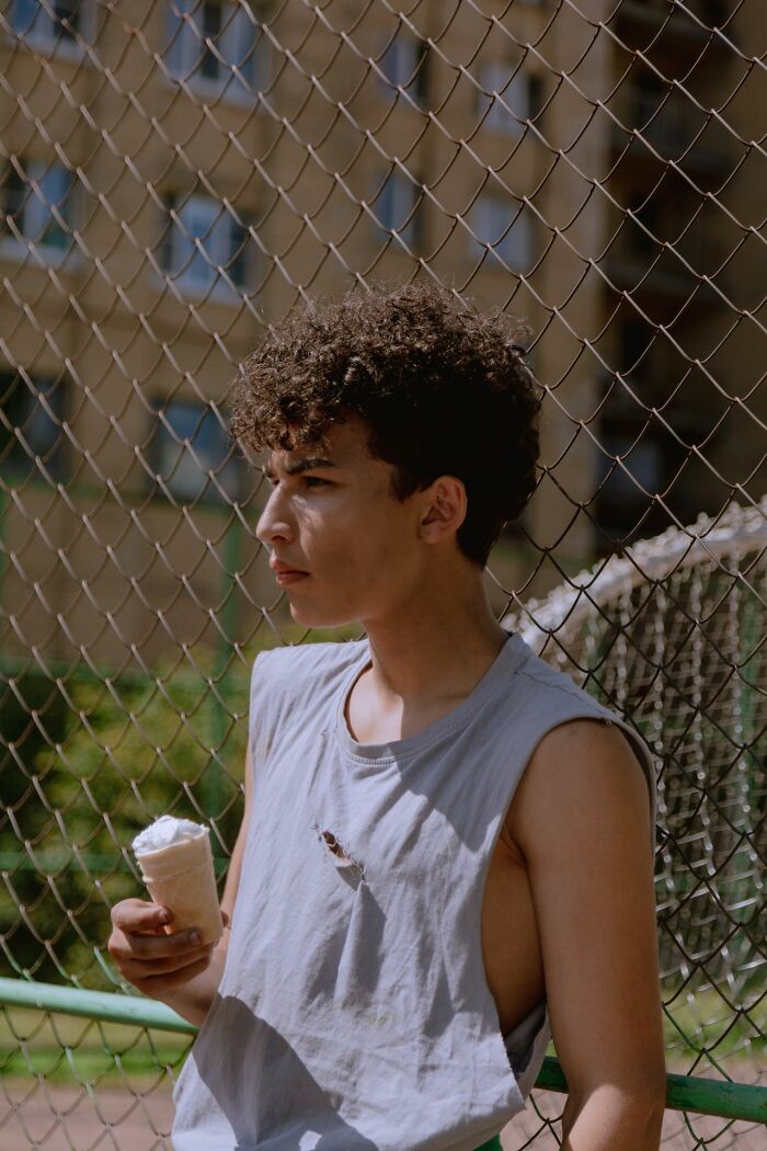 A young person in a t-shirt looks away, holding an ice cream cone by a chain-link fence, symbolizing simple joys.