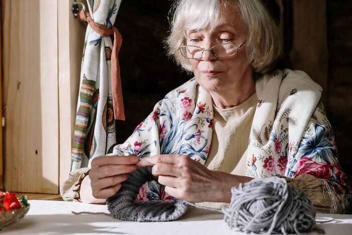 An elderly woman knitting, with a ball of yarn, possibly reflecting on something she overheard.
