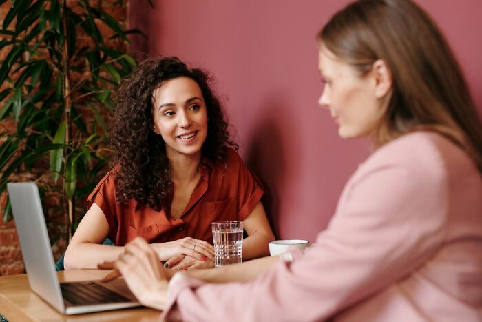 Two women conversing at a table with a laptop and a drink, enjoying hot tea or coffee.