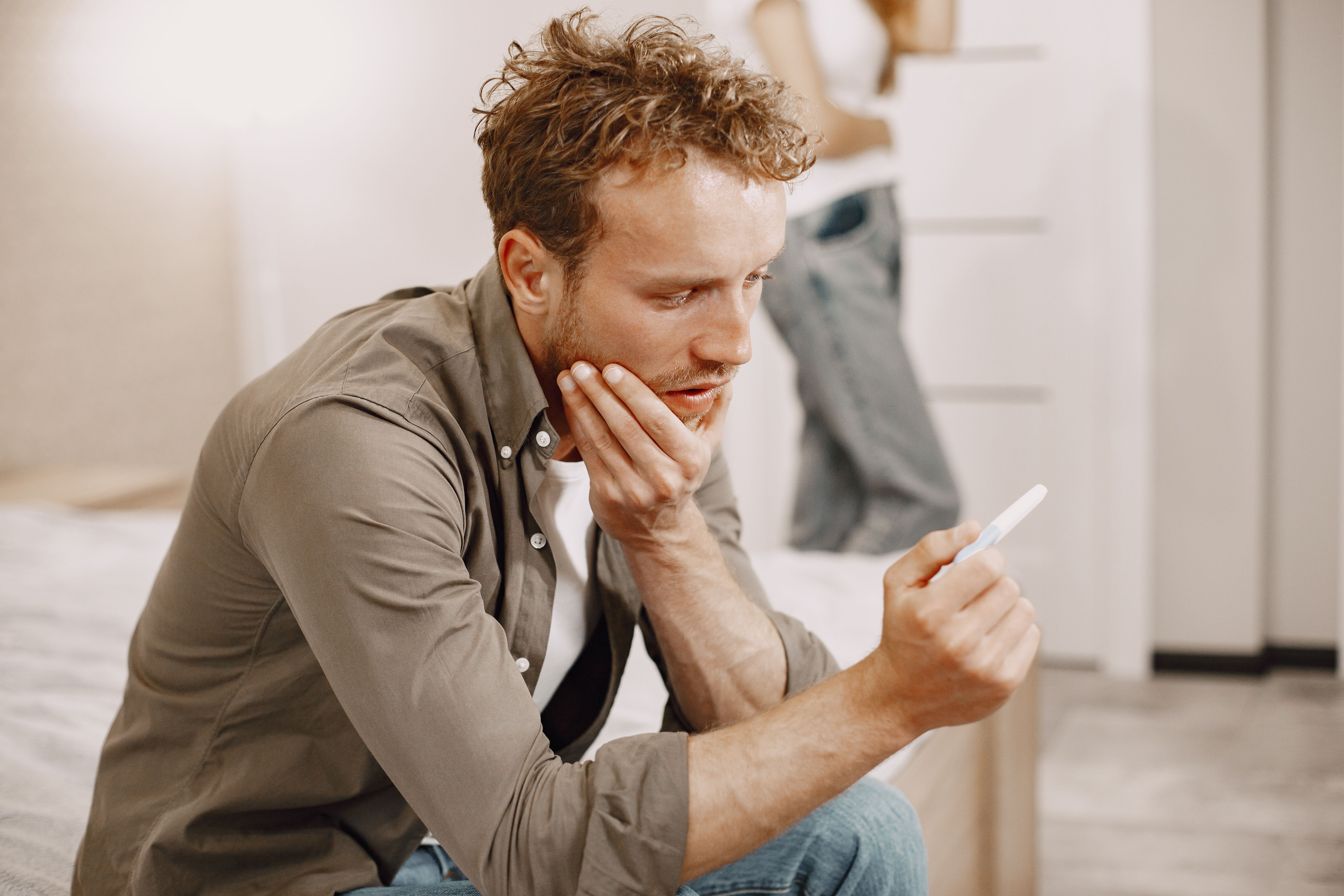 Worried man looking at pregnancy test in hand with woman blurred in background Worried man looking at pregnancy test in hand with woman blurred in background