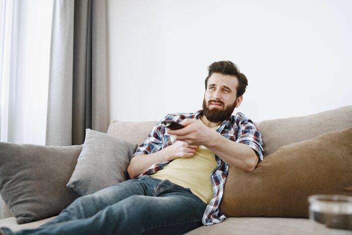 A man on a couch looking worried while holding a remote, showcasing dating red flags.
