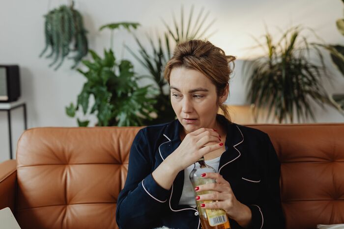 Woman on a brown sofa in dark blue pajamas, holding a glass bottle; depicting a nightmare airline seatmate story.
