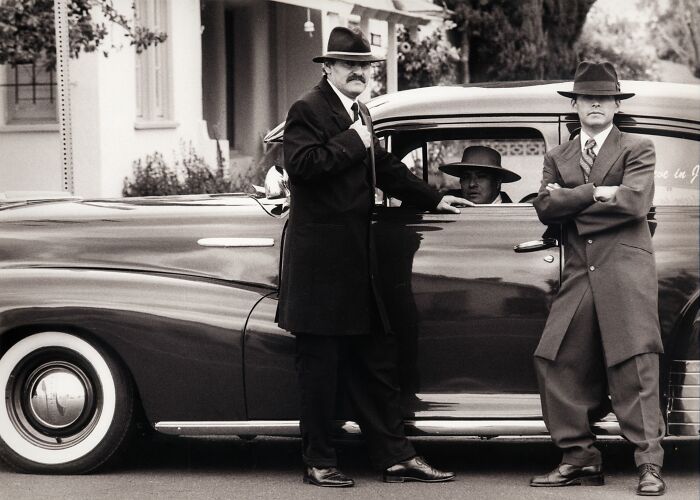 Three men in fedoras and suits standing by a classic car, hinting at secrets that came to light.