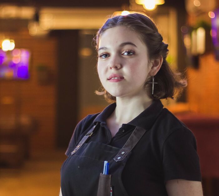 Young female restaurant server with apron standing in dining area