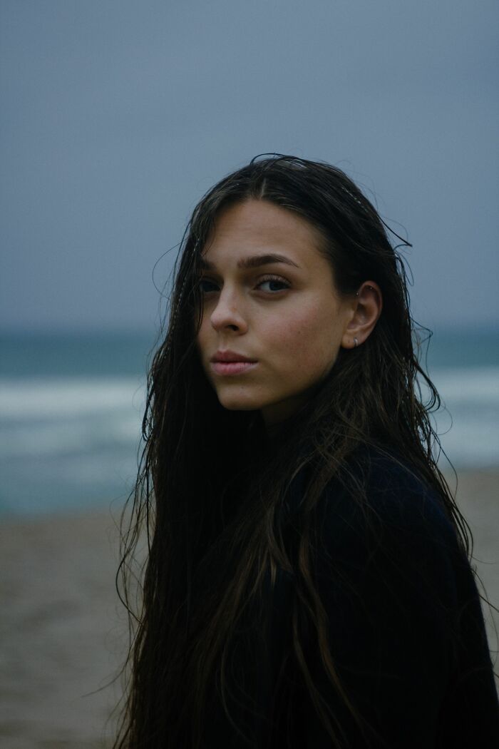 A woman with long wet hair stands by the ocean, her serious expression possibly reflecting on dating red flags.