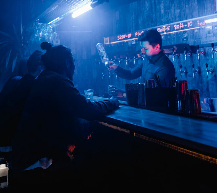 A bartender serves a customer at a bar, dimly lit with blue light, where people might overhear conversations.