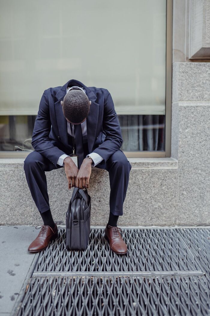 Businessman sitting on a ledge, head down, appearing exhausted and in need of hot tea.