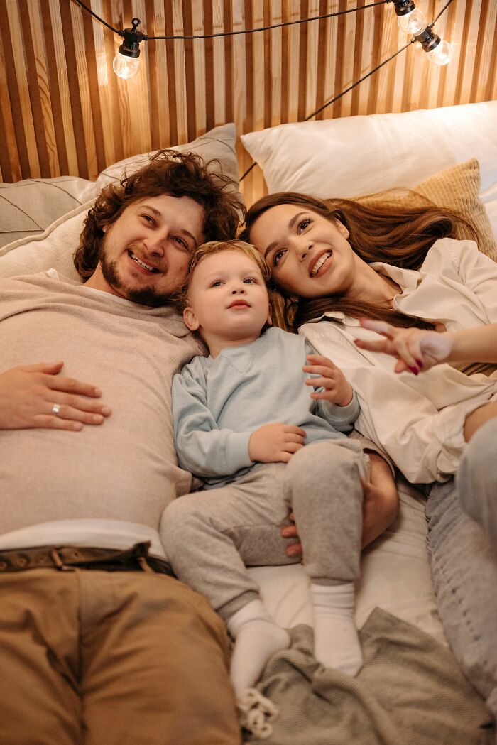 A happy family, a mother, father, and young child, lie together in bed, representing childhood beliefs about family.