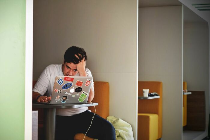 A person with their hand on their head, focused on a sticker-covered laptop, experiencing stress instead of simple joys.