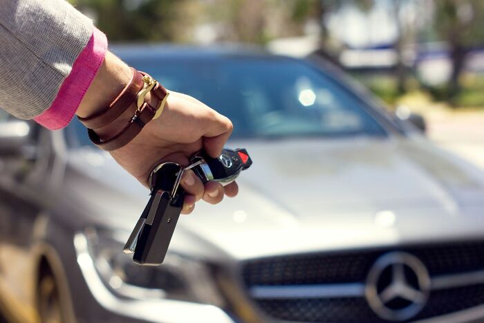 A hand holding car keys with a luxury car in the background, a safety tip for women living alone.