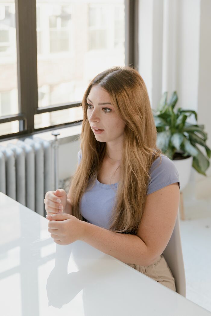 A young woman with long hair sitting at a table, looking thoughtful, as if she overheard something.