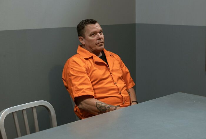 A man in an orange jumpsuit sits at a table, possibly depicting a sociopath during a therapy or interrogation.
