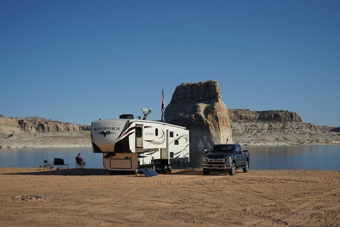 A fifth-wheel RV and a pickup truck parked on a sandy shore near water and large rock formations, revealing industry secrets.